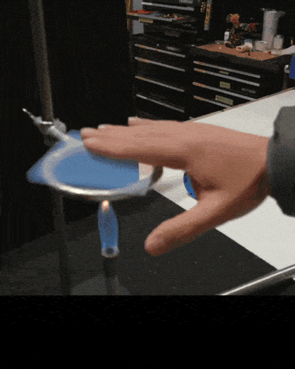 O mână trece rapid prin flacăra unui arzător bunsen albastru, prezentând un experiment științific. Fundalul prezintă echipamente de laborator și sertare.