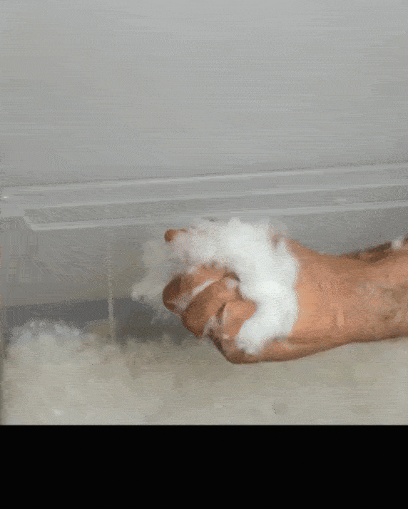 O mână zdrobește o grămadă de zăpadă sintetică pufoasă, permițându-i să cadă în cascadă pe o grămadă mai mare de dedesubt. Textura albă și moale sugerează o scenă de iarnă, deși este în interior. Fundalul este o suprafață simplă.