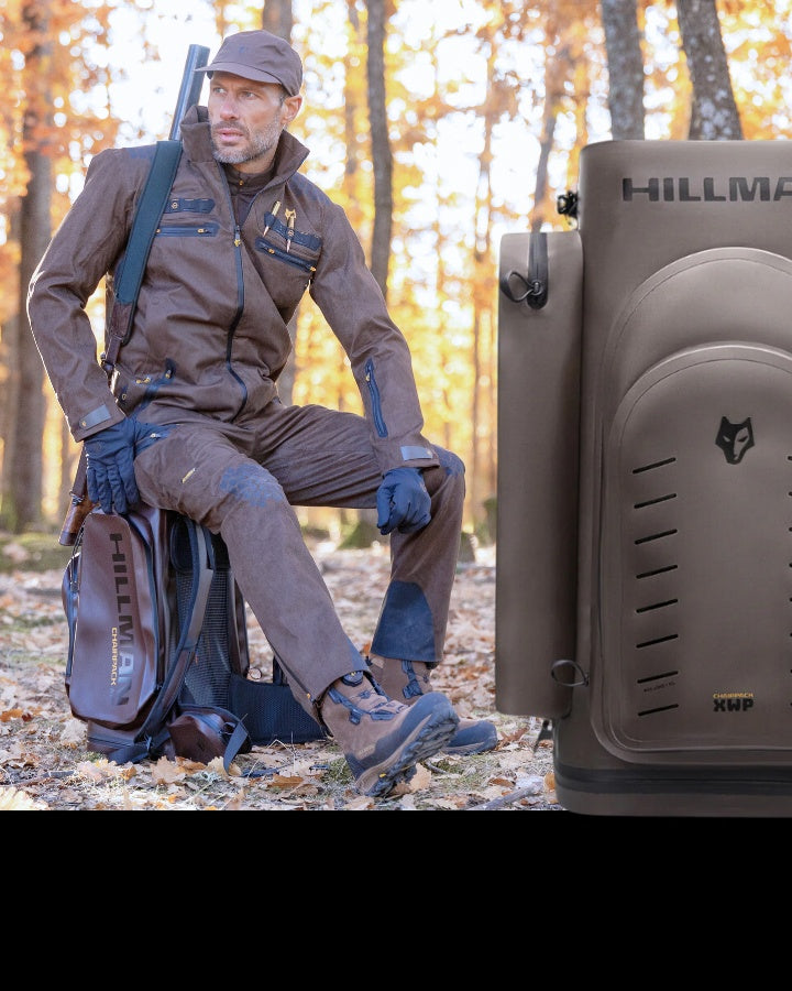 O persoană care poartă o ținută maro de exterior, cu șapcă și mănuși, stă ghemuită într-o pădure cu frunze de toamnă pe pământ. Ei poartă un rucsac și o pușcă. În partea dreaptă se află o cutie mare maro pentru echipament, cu un logo.