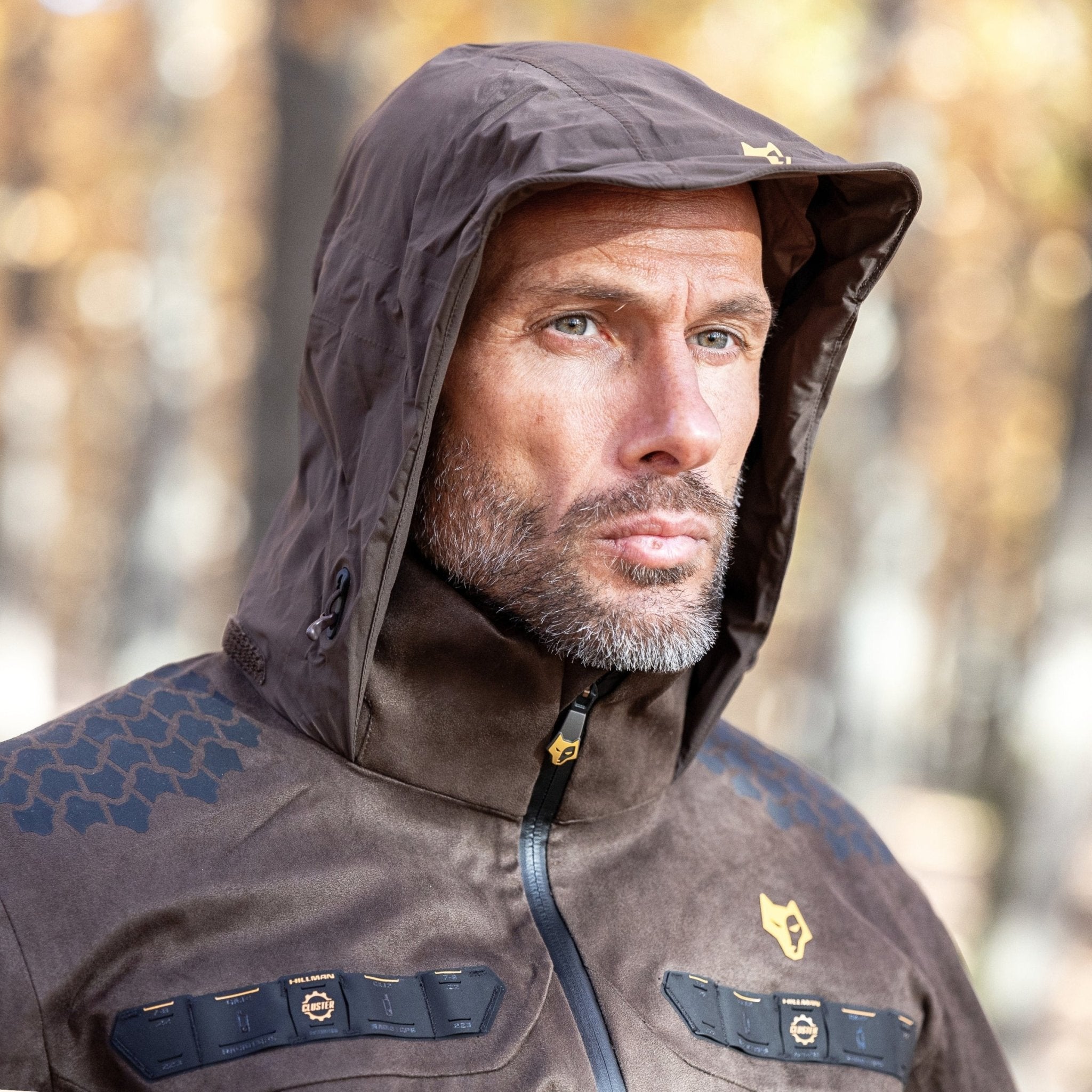 O persoană într-o jachetă maro Hillman geacă vânătoare ușoară 5WL stă în aer liber, privind în depărtare. Fundalul prezintă copaci de toamnă încețoșați. Această jachetă impermeabilă prezintă modele și embleme distinctive.